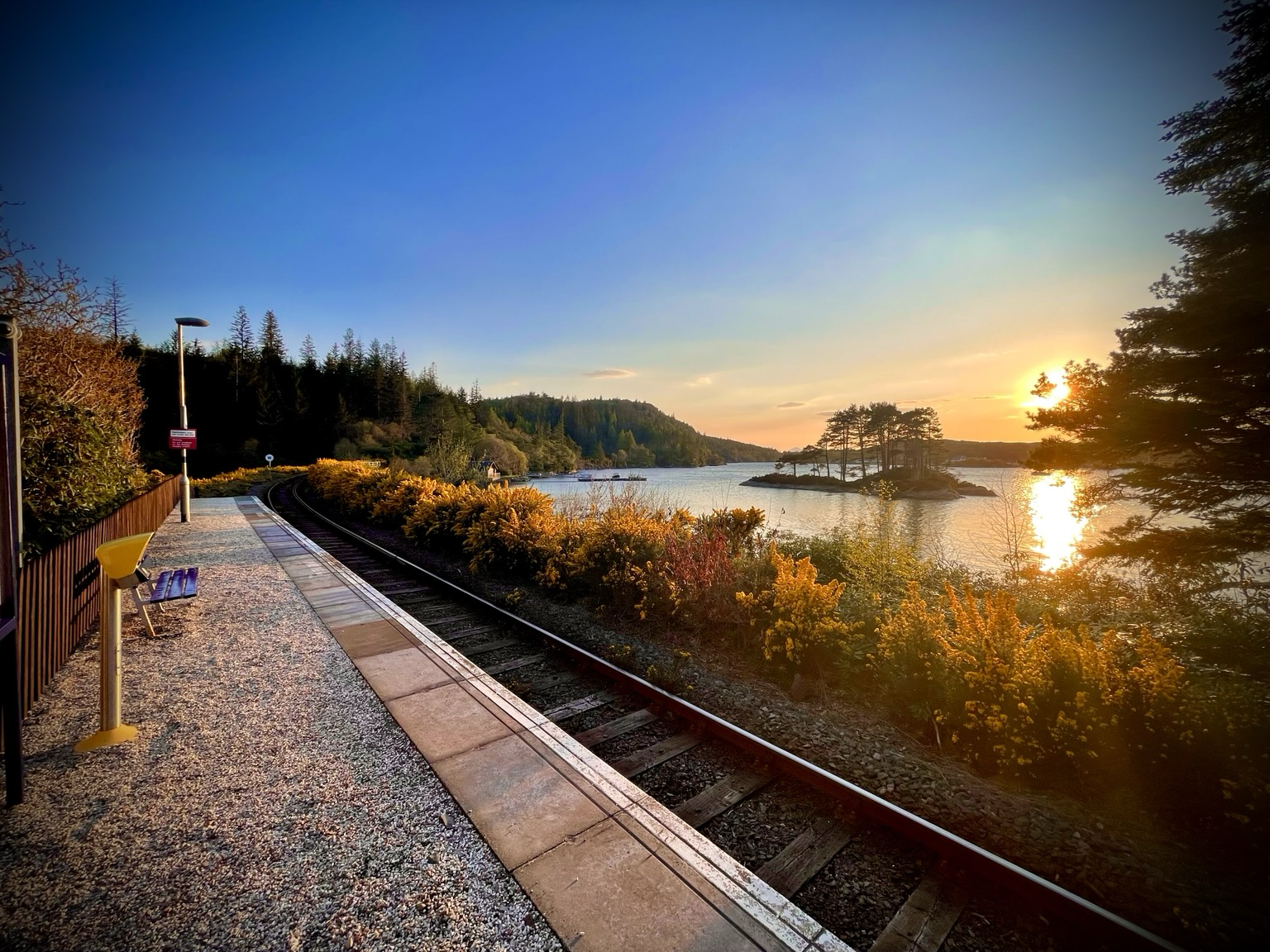 A very serene Duncraig railway station looking over the water to the village of Plockton in the Scottish Highlands. The end of a beautiful summer’s evening with the sun setting with an orange glow in the sky. The station itself is a request stop on the railway line between Inverness and Kyle of Lochalsh, where to board a passing train, you simply stick out your arm to wave it down as you might a taxi.