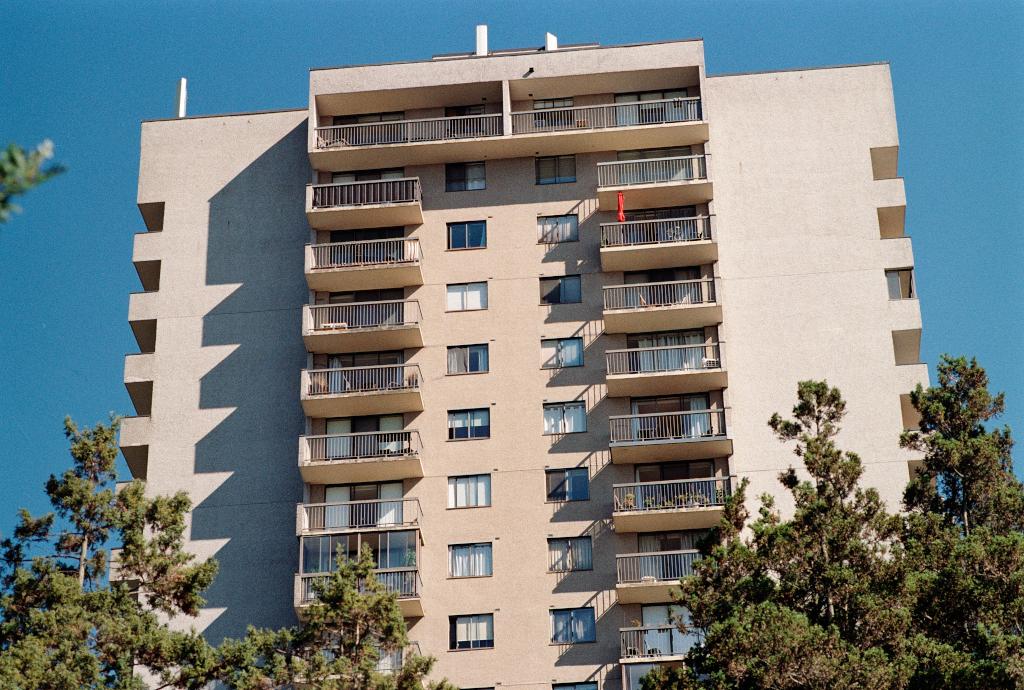 The upper stories of a rather boring apartment building of sand and brown colours. One one balcony a folded red umbrella stands as if in protest.