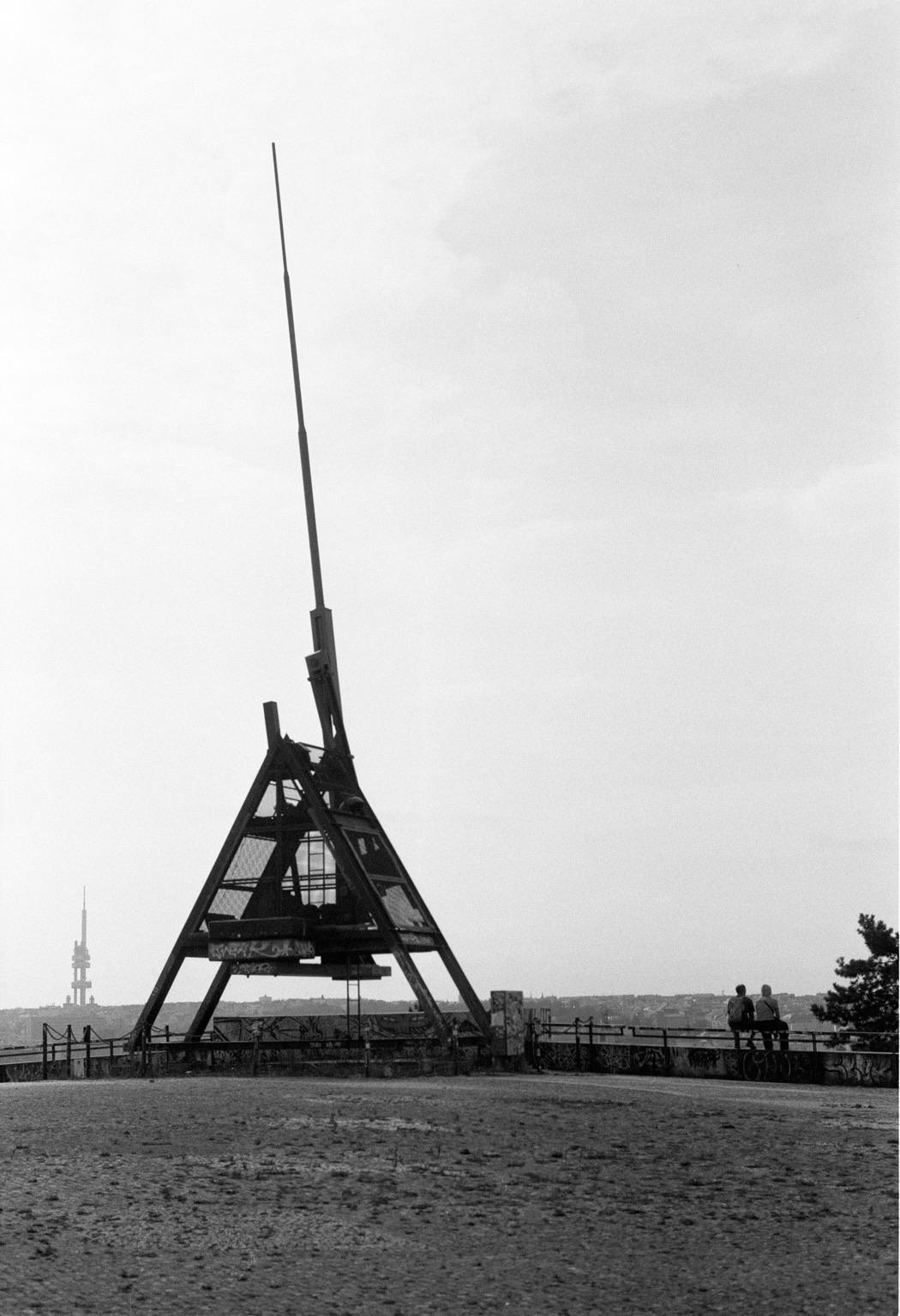 A black and white image featuring a large triangular structure with a tall antenna atop a hill. Two people are seated on a nearby railing, overlooking a landscape in the background. The sky appears overcast, contributing to the muted atmosphere.