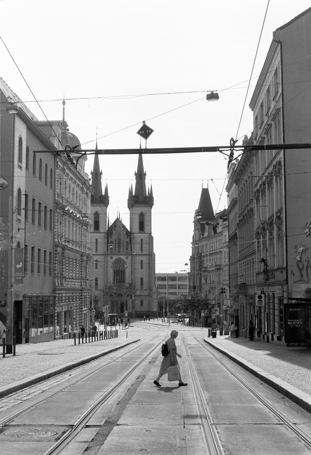 A black-and-white photo of a street with tram tracks, showing a person walking across the road. Historic buildings line both sides, with a church featuring tall spires in the background. The scene is bright and captures an urban atmosphere.