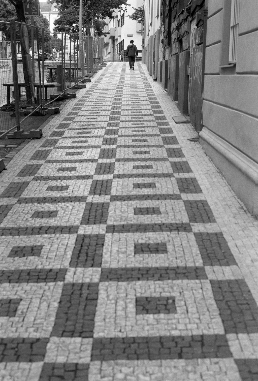 A person walking down a narrow, patterned cobblestone sidewalk. The path features a checkerboard design in shades of gray and black. Nearby, there are construction barriers, and buildings line the street. The scene is captured in black and white.