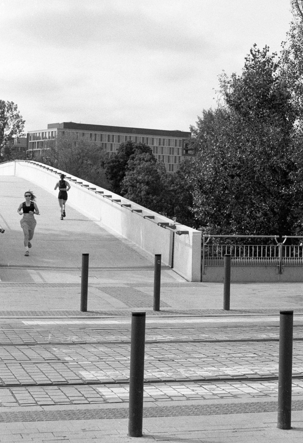 A black and white image of two joggers running on a curved bridge. In the background, there are trees and a modern building. The foreground features a textured pavement with several poles and a tram track.