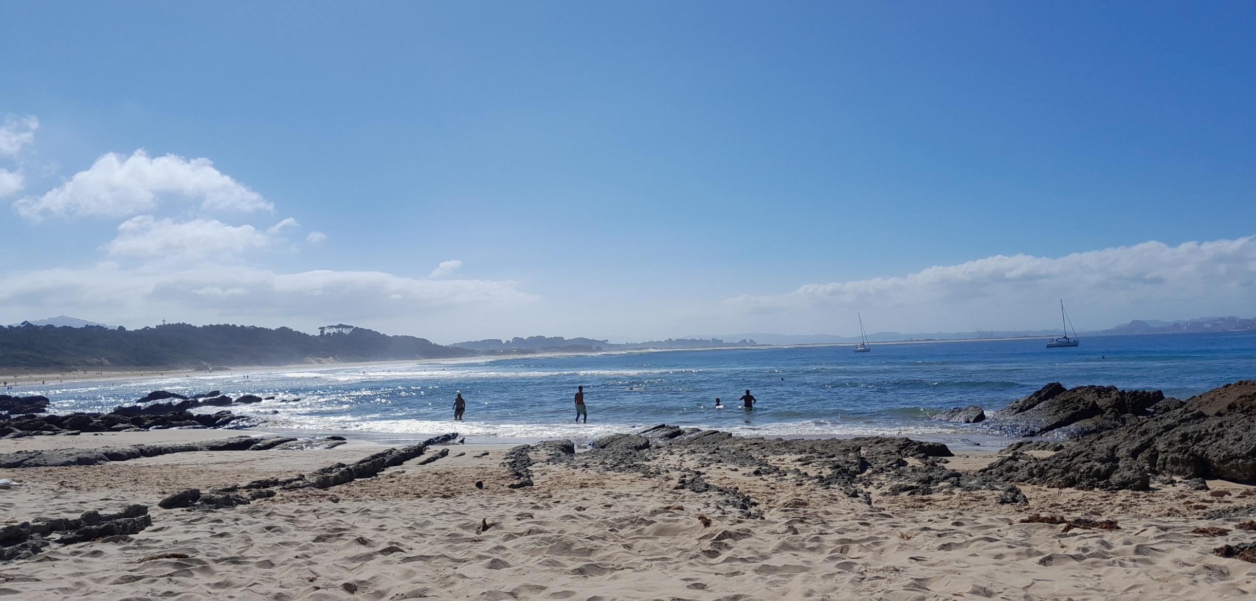 La playa, rocas negras en la orilla, el mar azul oscuro con olas espumosas, algo de gente pero no mucha, y al fondo, más playa, unas nubes bajas, un par de barcos y montañas al final