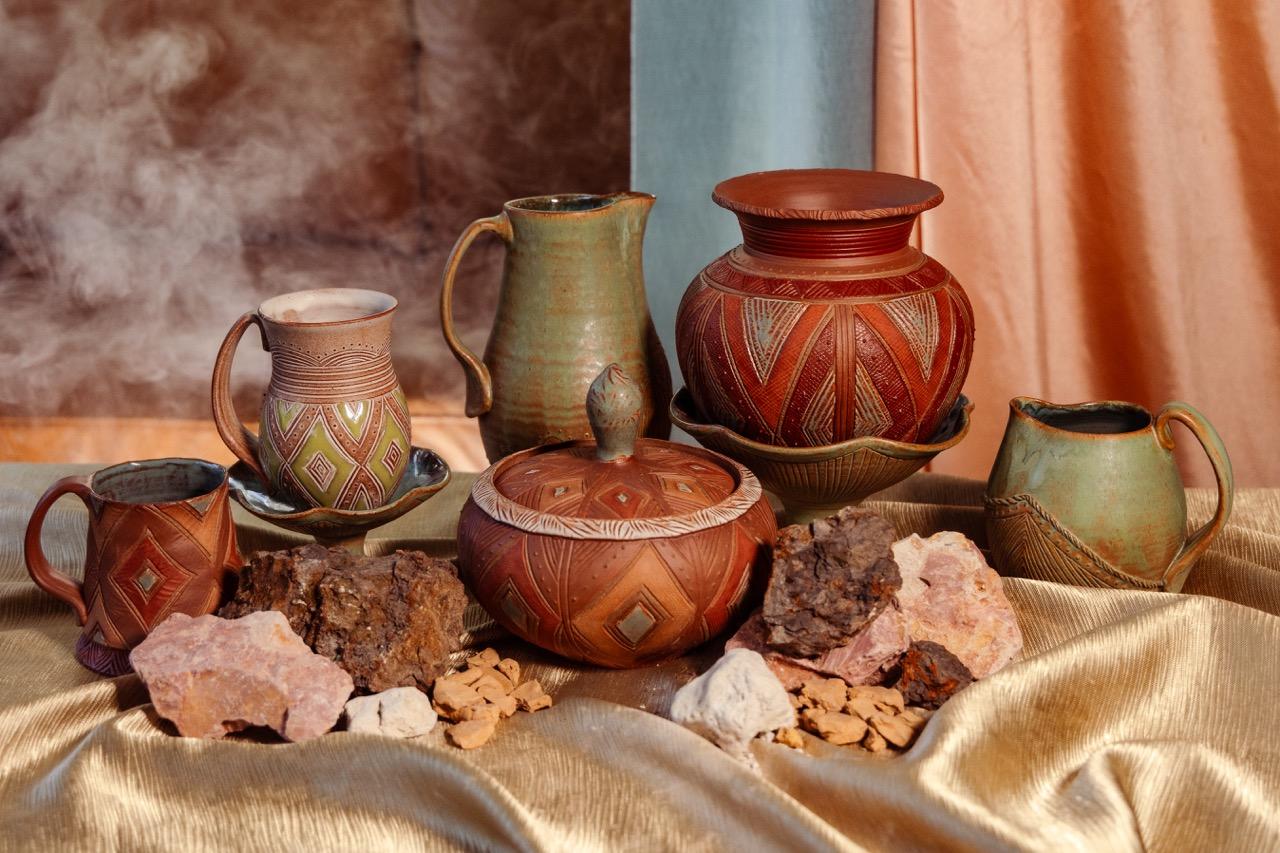 Several pieces of pottery and multi-colored rocks of clay arranged on top of a table covered in gold fabric. The pottery features decorative geometric patterning and warm earth tones with light green and light blue glazes that are sheer enough to allow the undertones of red clay to come through the surface. There are peach and blue curtains in the background to the right and a small cloud from a fog machine in the background to the left. The overall look of the photo is similar to a classical French still life painting.
