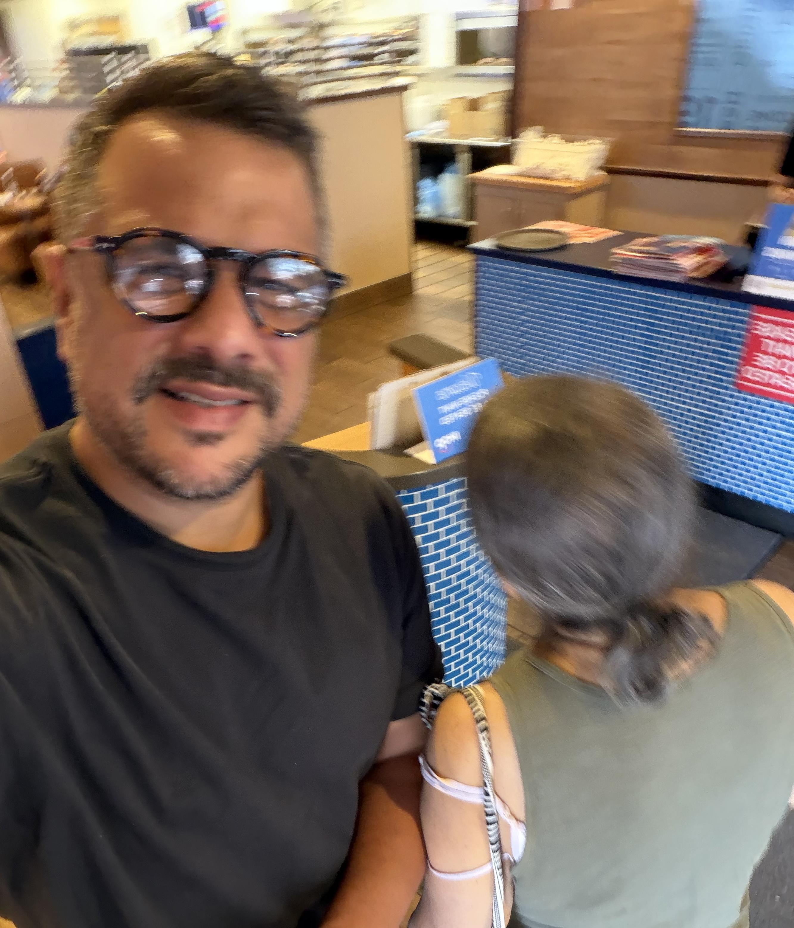 Blurry photo of man with glasses (me) taking a selfie in an unopened restaurant with a woman (mom) facing the opposite way, you can only see the back of her head. 
