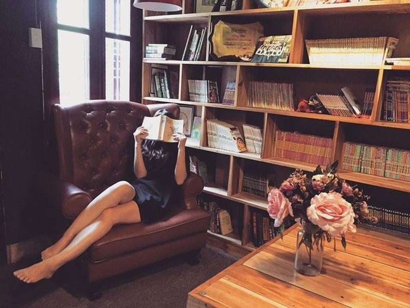 In this photo, a young woman is reading a book while sitting sideways on a large brown leather armchair next to a tall wooden bookshelf. Her legs are stretched out and crossed, and she is wearing a short black dress. Her face is hidden behind the book. The chair is a classic brown leather wingback, and the light wooden bookshelf is filled with books, comics, and manga.
