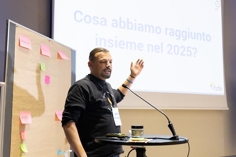 A speaker presents in a seminar room at SFSCON, standing beside a board covered with colourful sticky notes. A presentation slide in the background reads “Cosa abbiamo raggiunto insieme nel 2025?” (“What did we achieve together in 2025?”).