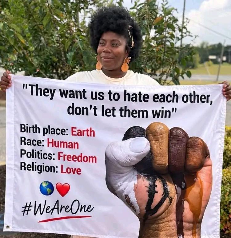 A person stands outdoors holding a white banner with diverse hands forming a united fist. The banner promotes peace, equality and unity with the words “They want us to hate each other, don’t let them win,” and highlights Earth as our birthplace, humans as our race, freedom as politics, and love as religion. The message ends with “#WeAreOne.” 🌍❤️
