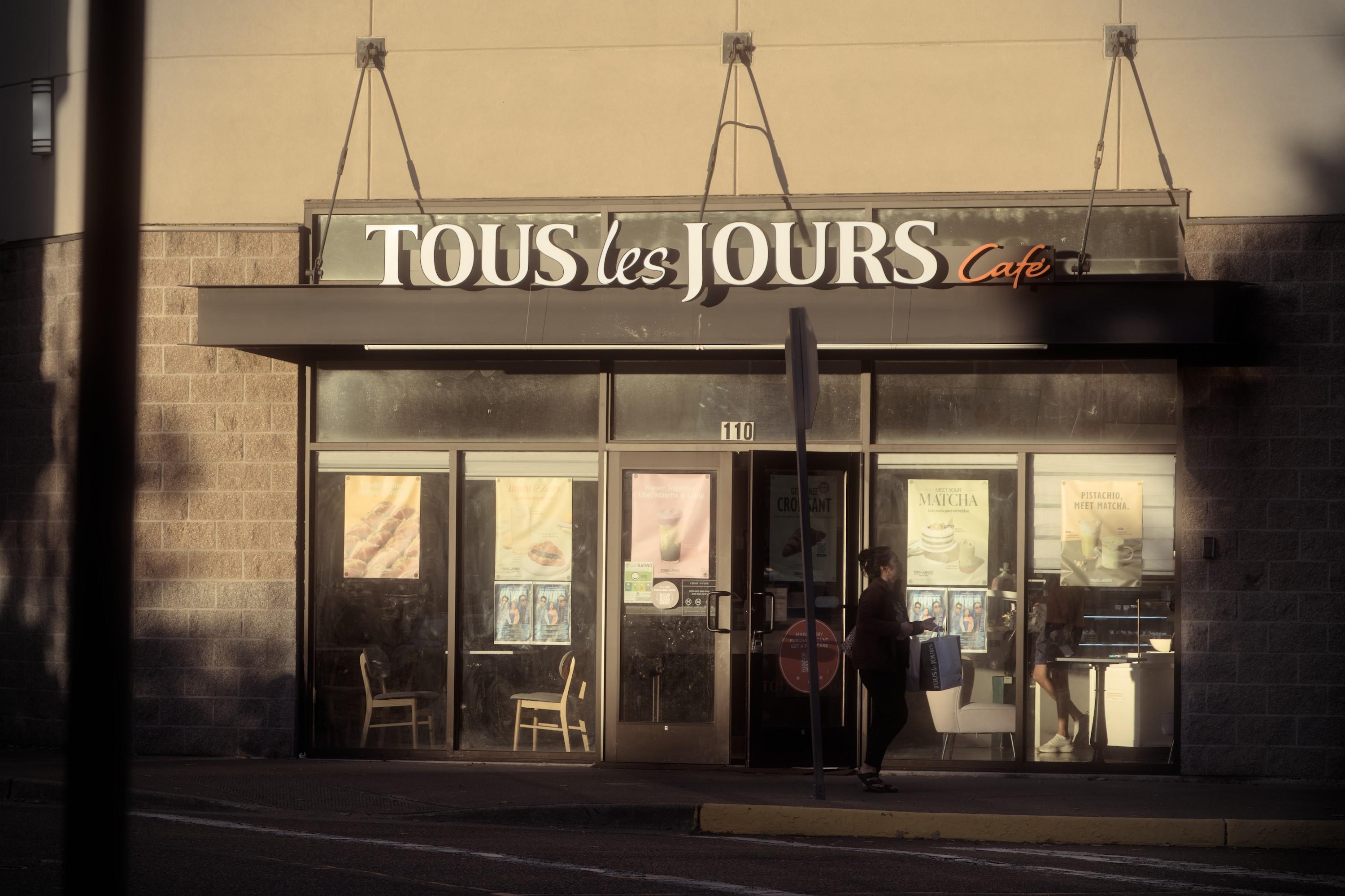 An entrance to the bakery store with a sign Tous Les Jours Cafe above it. The store is washed out in golden-tan tones of the setting sun, partially covered by deep shadows from trees and buildings. There's a drak silhouette of a person waking out from the store.