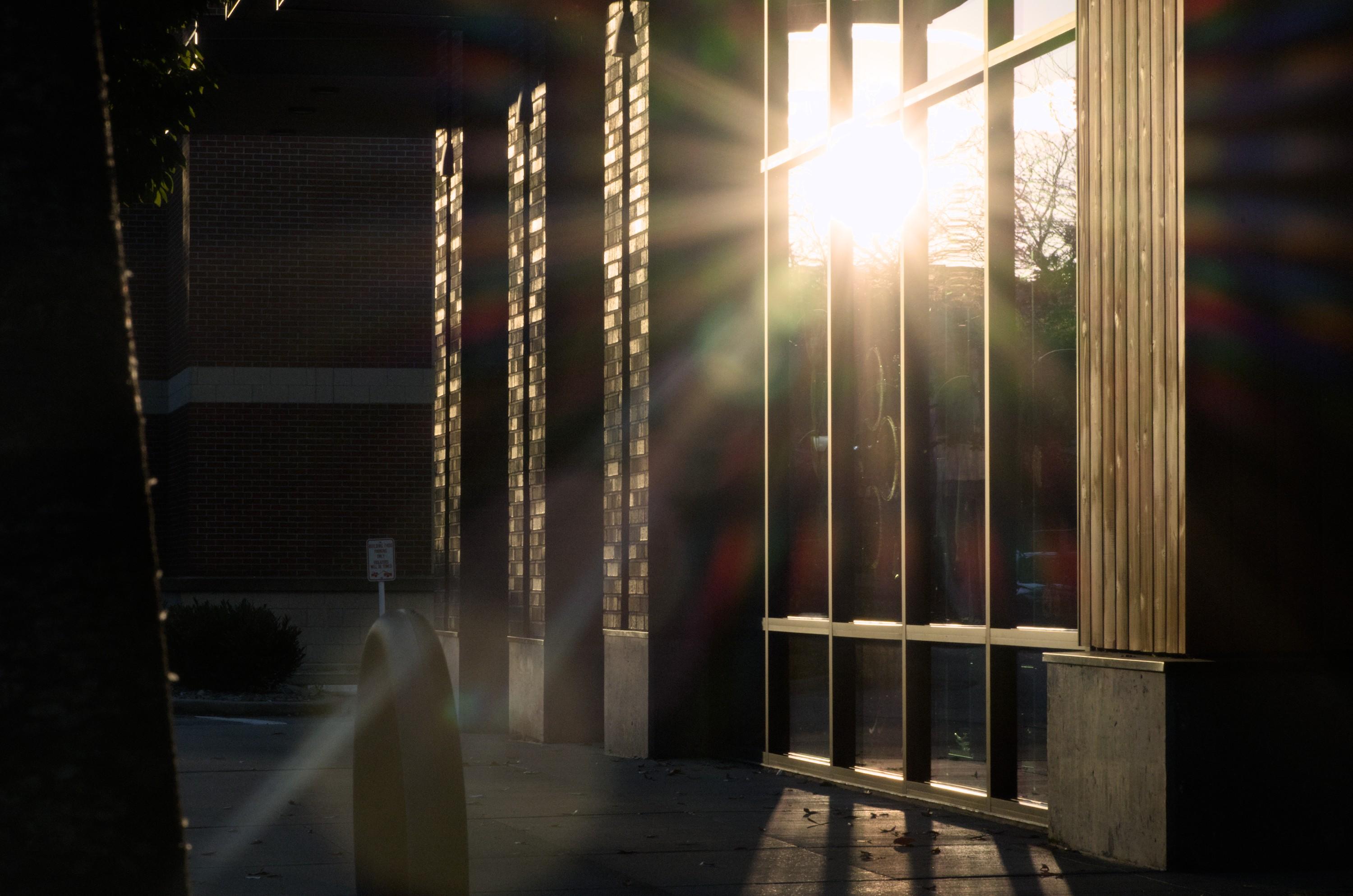 Reflection of the setting sun in a floor-to-ceiling window pane facing a sidewalk. The scene is overall dark, but the sun's reflection is a bright spot with separate rays of light going in all directions, along with other difraction effects due to the camera lens.