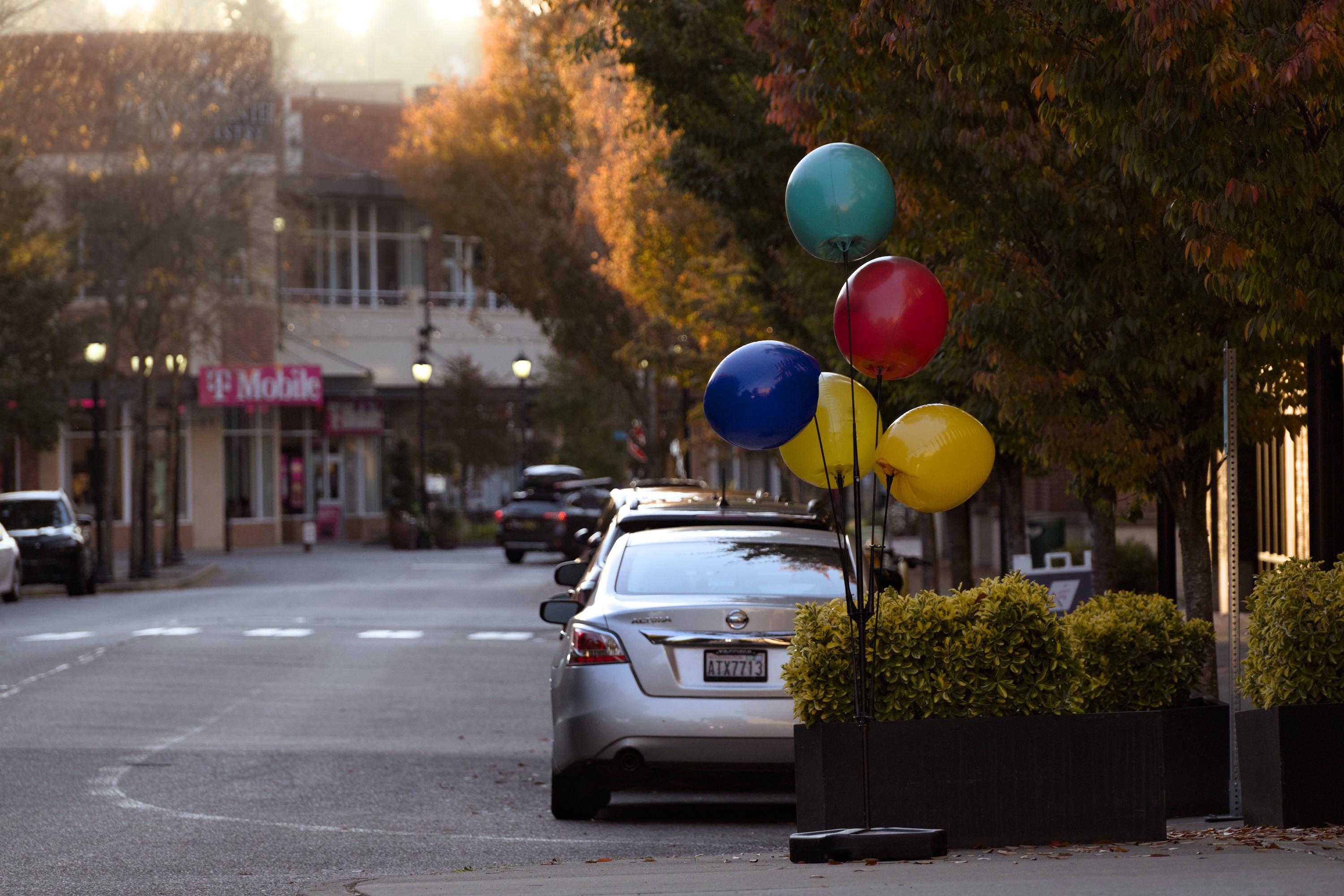 A look down a deserted city street with the right half of the frame occupied by trees with canopies in fall colors, dark green to yellow. In the foreground there's a tall stand with a bunch of colorful balloons standing on the sidewalk, and the back of a silver-colored parked car. There is a very vright patch of sky above the buildings in the background giving the contrast to the twilit street.