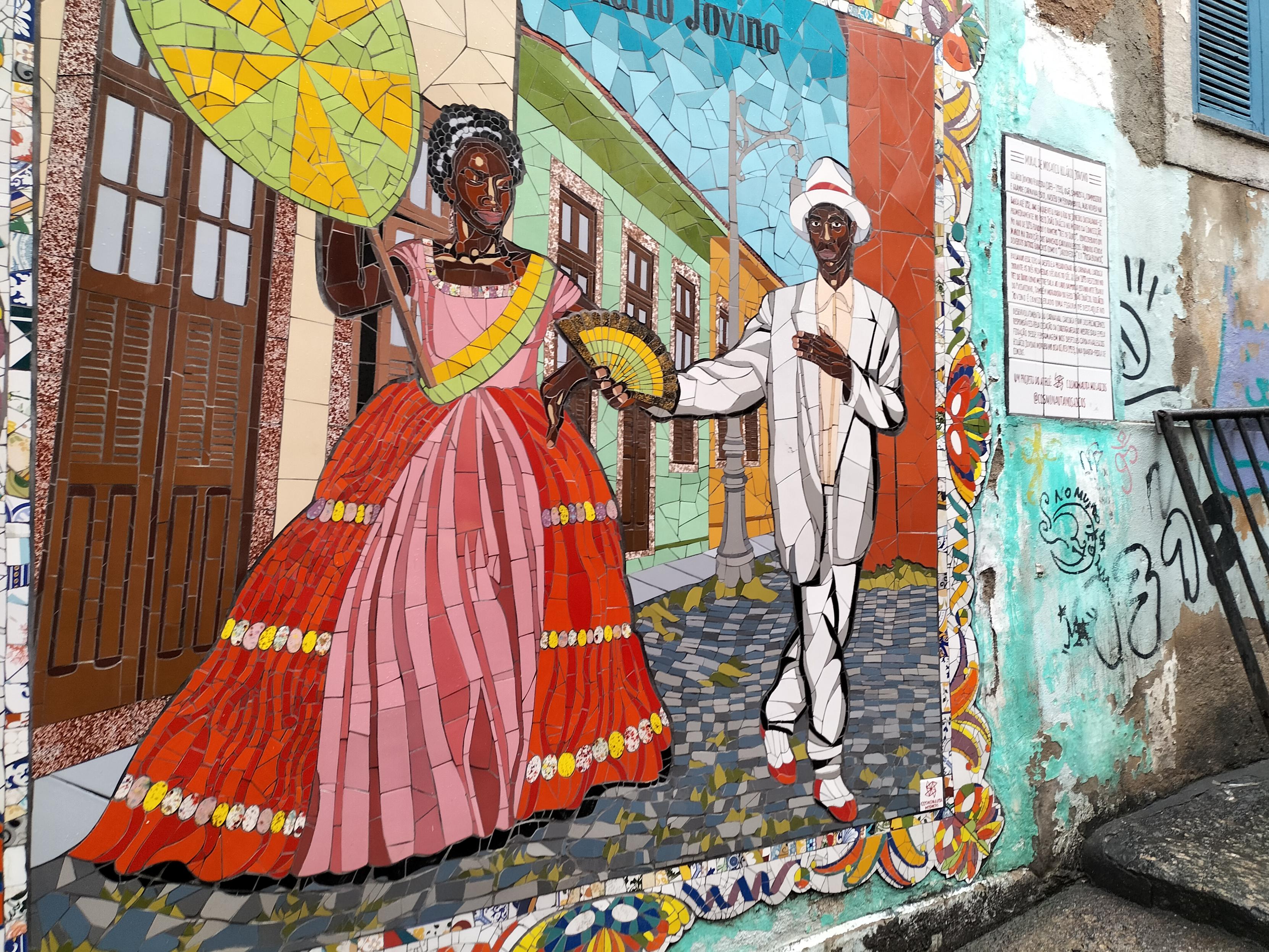 Resistencias africanas.Mosaico de mujer negra  en traje de época estilo "colonial"colorido y hombre negro en traje blanco de la misma época..Letra negra sobre cielo azul que pone Jovino (explicación que no se lee sobre importancia musical de ese hombre en la  historia de la Samba y el carnaval como maestro musical)