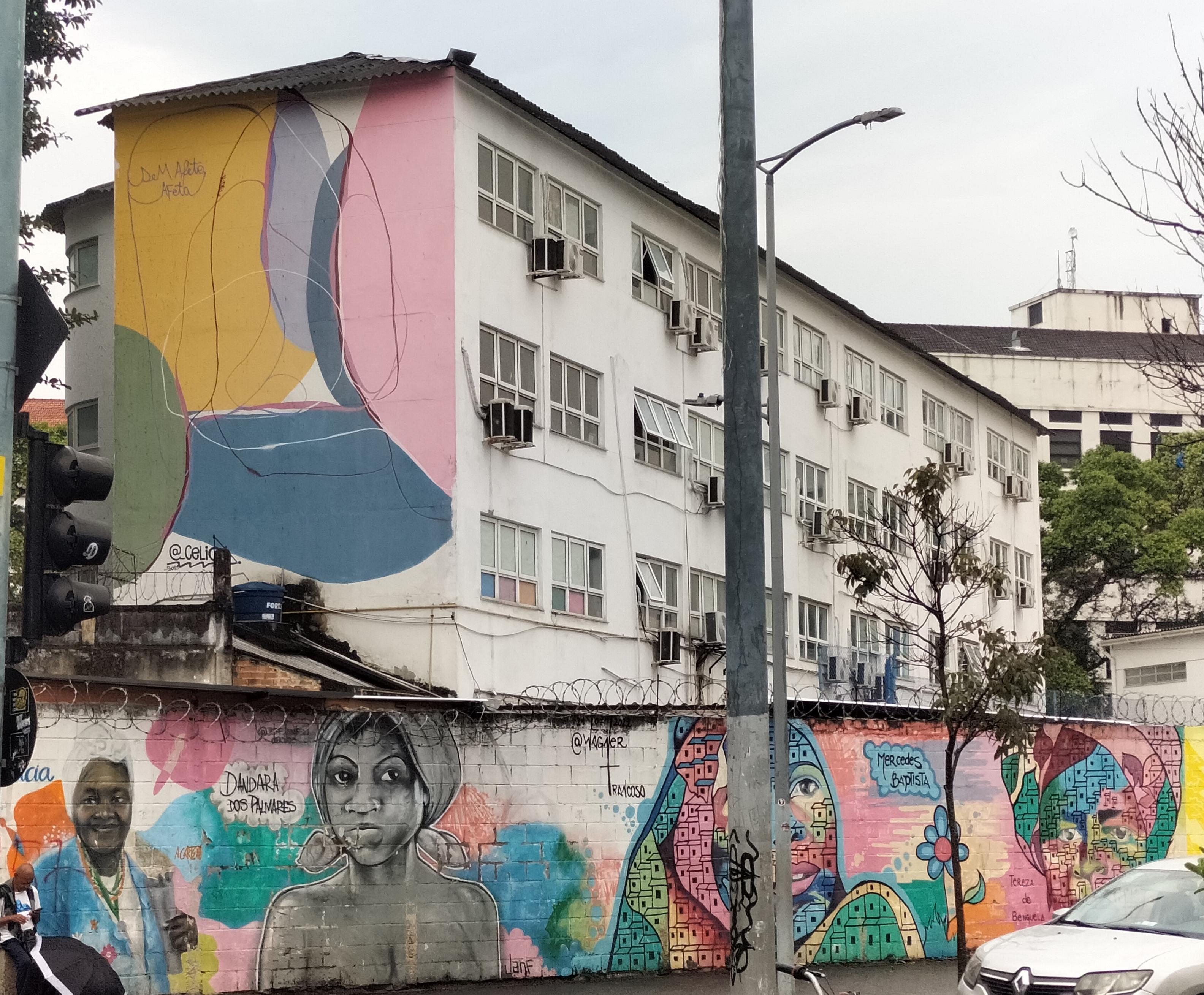 Mural de formas abstractas coloridas en una esquina de edificios. Debajo,muro con una mujer negra mayor en ropa de calle,otra joven en busto con pañuelo e insinuación de pecho,otra joven abstracta con colores variados y edificios en su interior y otra similar pero mayor tapada por un coche