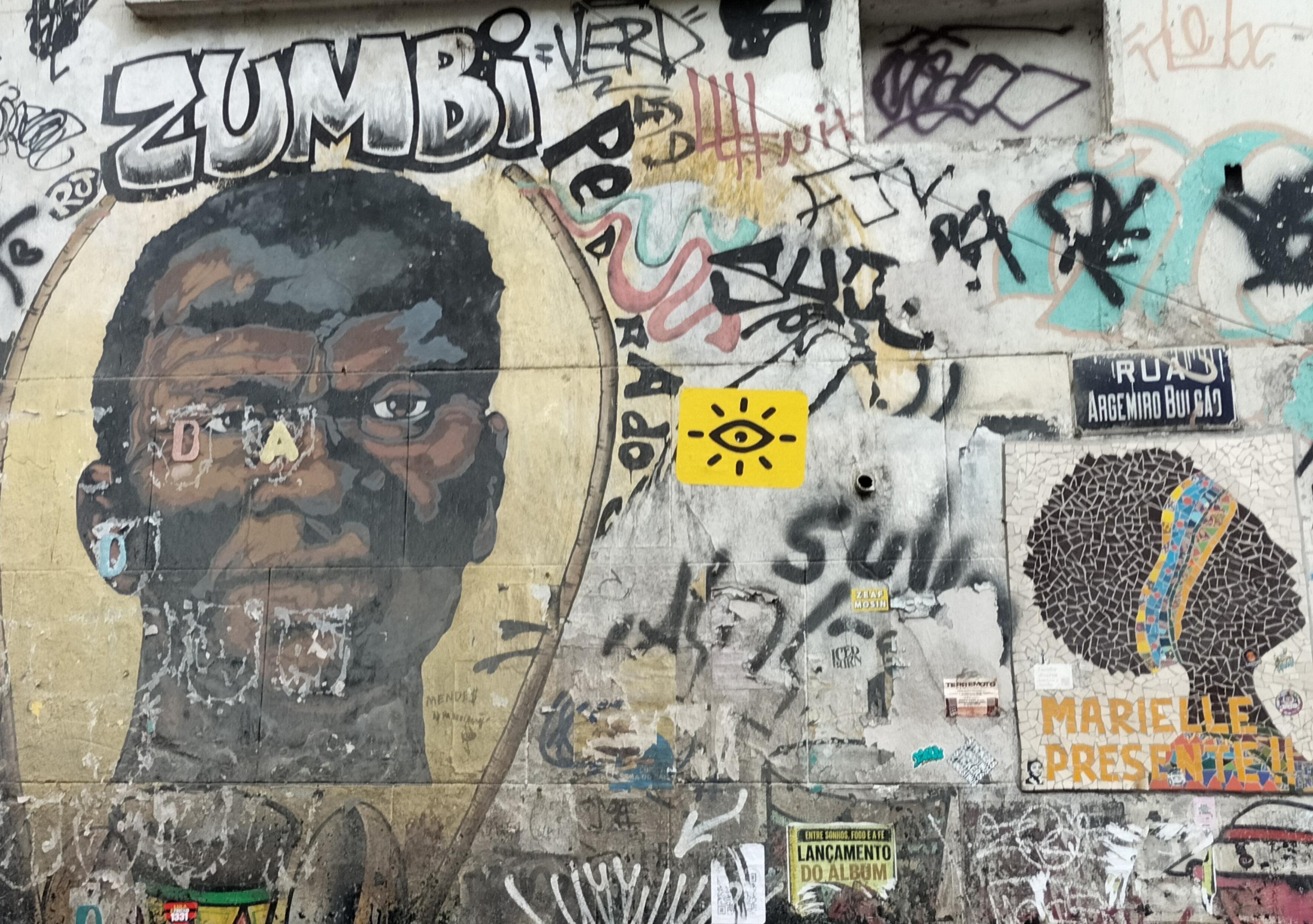 Retrato de hombre negro con letras grandes que pone Zumbi y mosaico de mujer negra de perfil con diadema de colores amarillos y azules y letras amarillas Marielle Presente!!