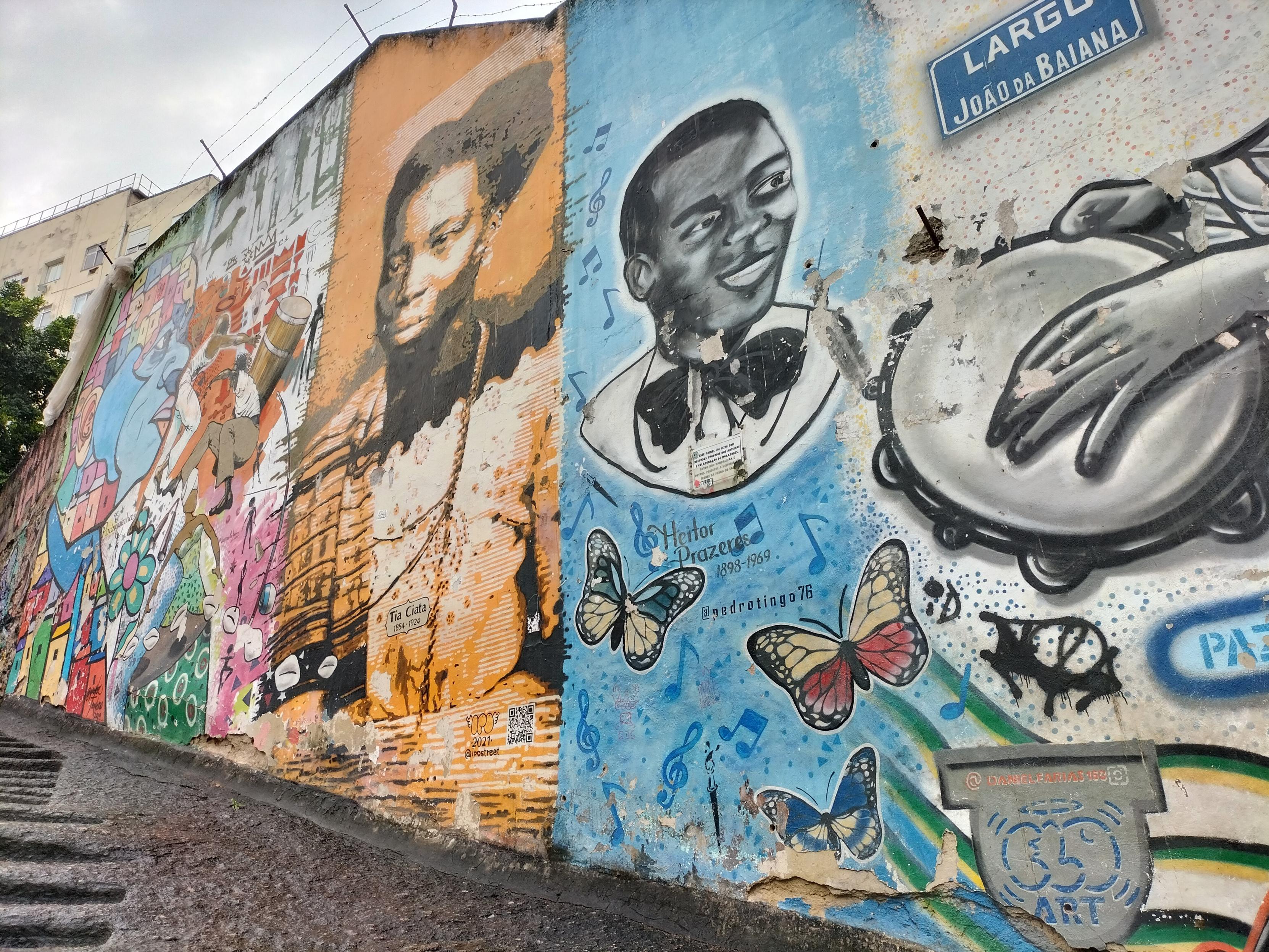 Conjunto de grafitis bajando Pedra do Sal.Figura colorida que no se distingue bien.Grafiti realista de mujer negra en naranjas con cartelito Tia Ciata 1854-1924.Retrato de hombre negro con nombre Heitor Prazeres 1898-1969, fondo azul con notas musicales y mariposas con firma de autoría @pedrotingo76 y mano tocando pandereta en blanco y negro.