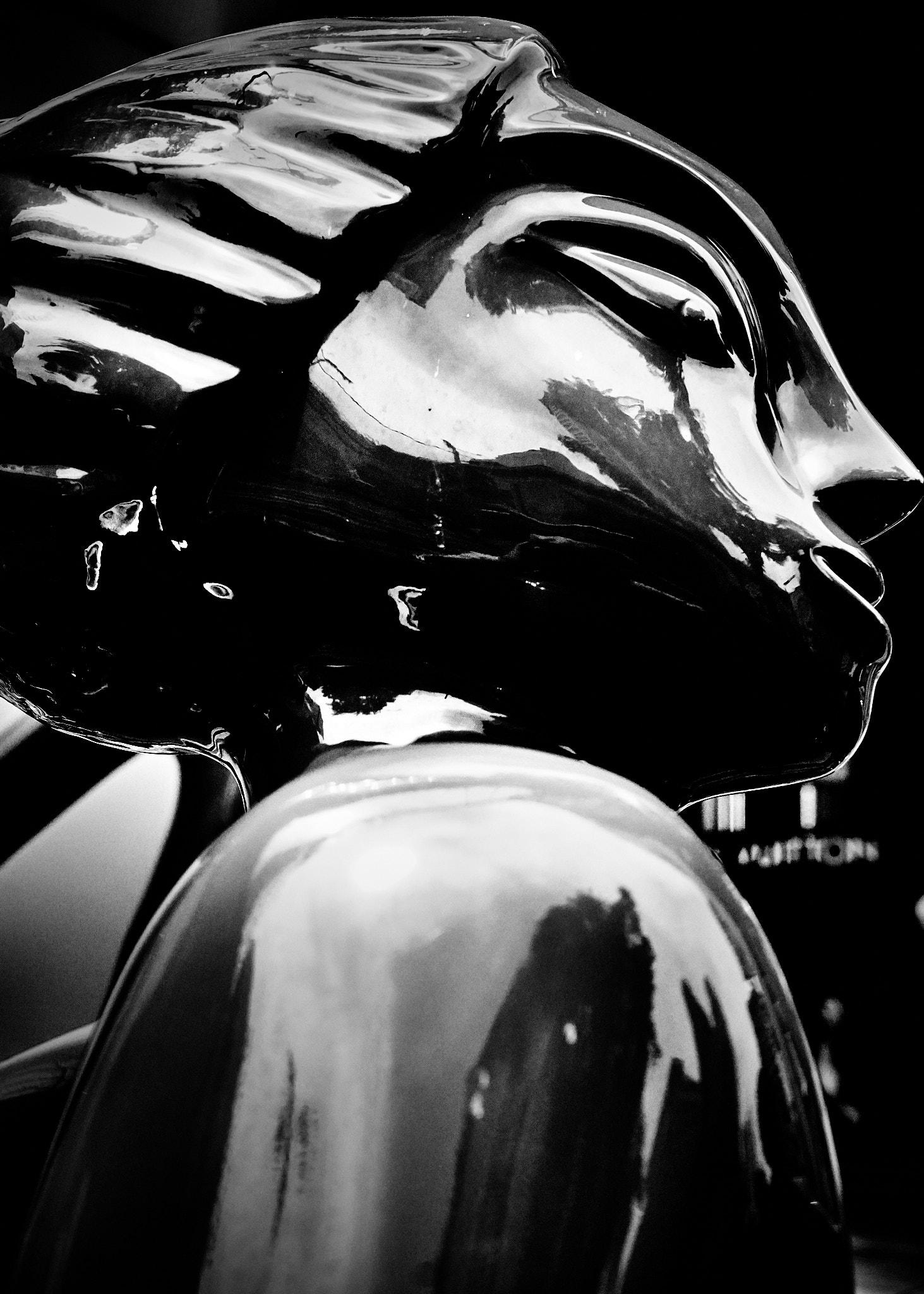 A close-up view of a shiny black sculpture resembling a stylized human face in profile. The surface reflects light and nearby objects, creating a glossy appearance. The background is dark, with hints of text and shapes that are not clearly defined.