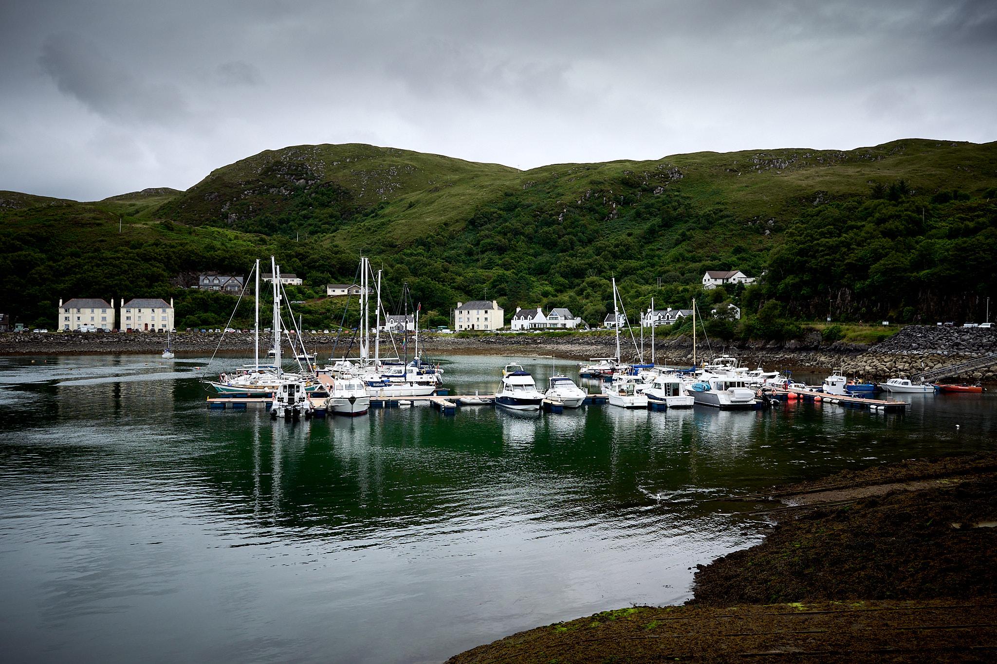 A harbor scene featuring multiple boats docked in calm water. In the background, green hills rise, with several buildings visible, including white and gray structures. The sky is overcast, indicating a cloudy day.