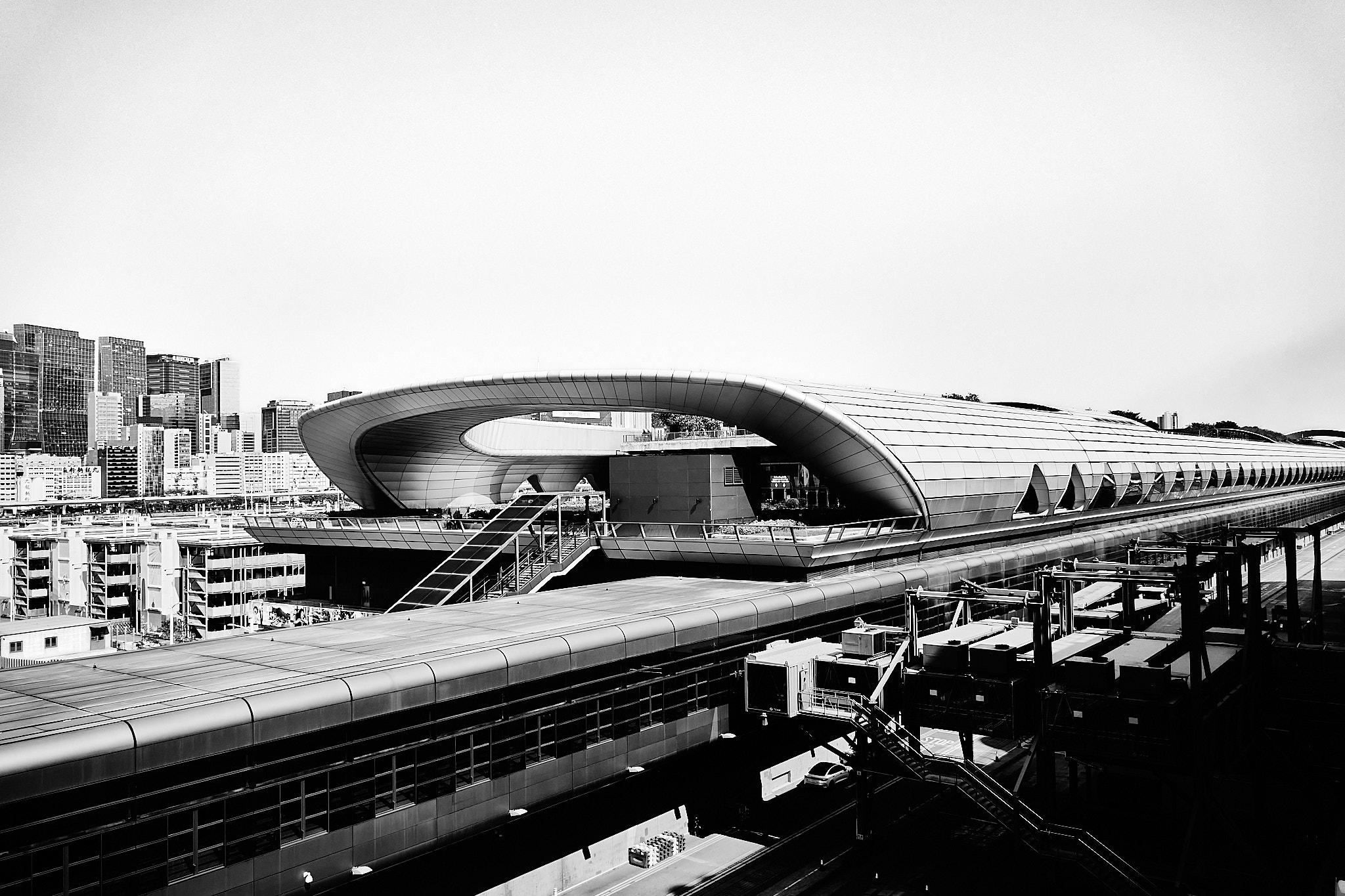 A modern architectural structure with a curved roof is prominently displayed. The foreground features a flat roof and a series of stairs leading up to the main building. In the background, a city skyline with tall buildings is visible, set against a clear sky. The image is in black and white.