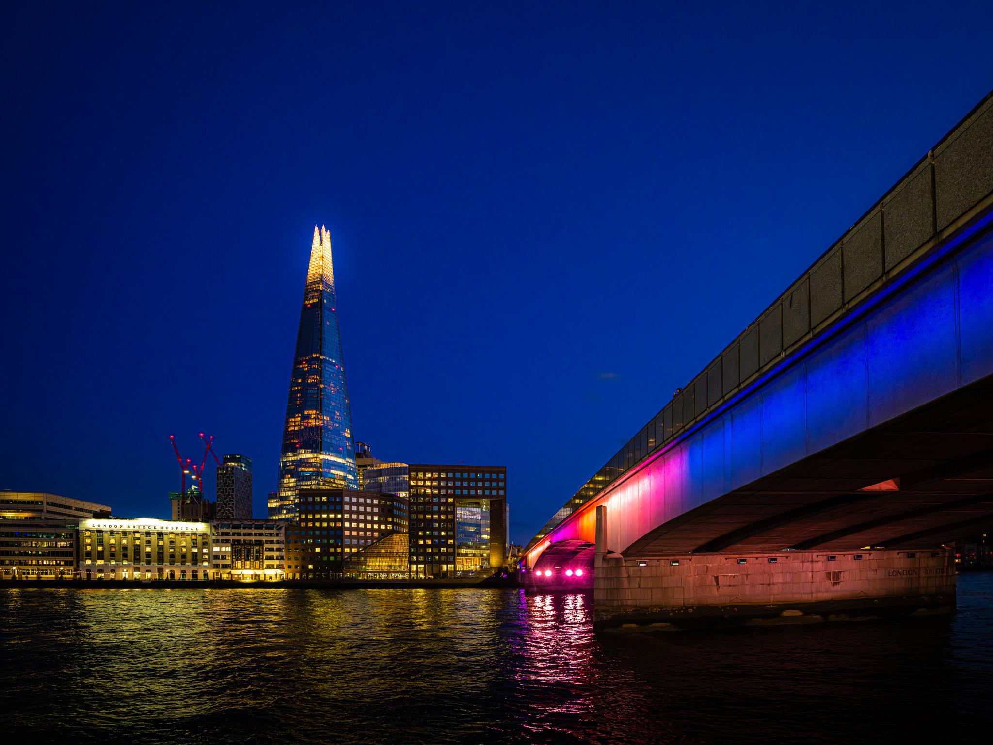 A wide night-time cityscape of London under a clear, very dark blue sky, taken by European elopement and wedding photographer Sturmsucht. The pale glow of a low cloud or haze sits just above the horizon on the right.

Right of centre, The Shard dominates: a glass pyramid tapering to a bright, pointillist-lit spire, its body speckled with warm yellow and orange office lights and a scatter of red warning lights. Around it cluster lower modern blocks — one with a prominent grid of lit windows, another showing the word "news" on a lit sign — while to its left the pale stone facade of London Bridge Hospital glows warm yellow, its name spelled out in lit letters along the roofline. Behind it, the red lattice of two construction cranes and a dark mid-rise building with a perforated facade stand against the sky, and further left a lower white building row extends off-frame.

Sweeping diagonally in from the upper right and filling the right third of the frame is a large concrete road bridge — i