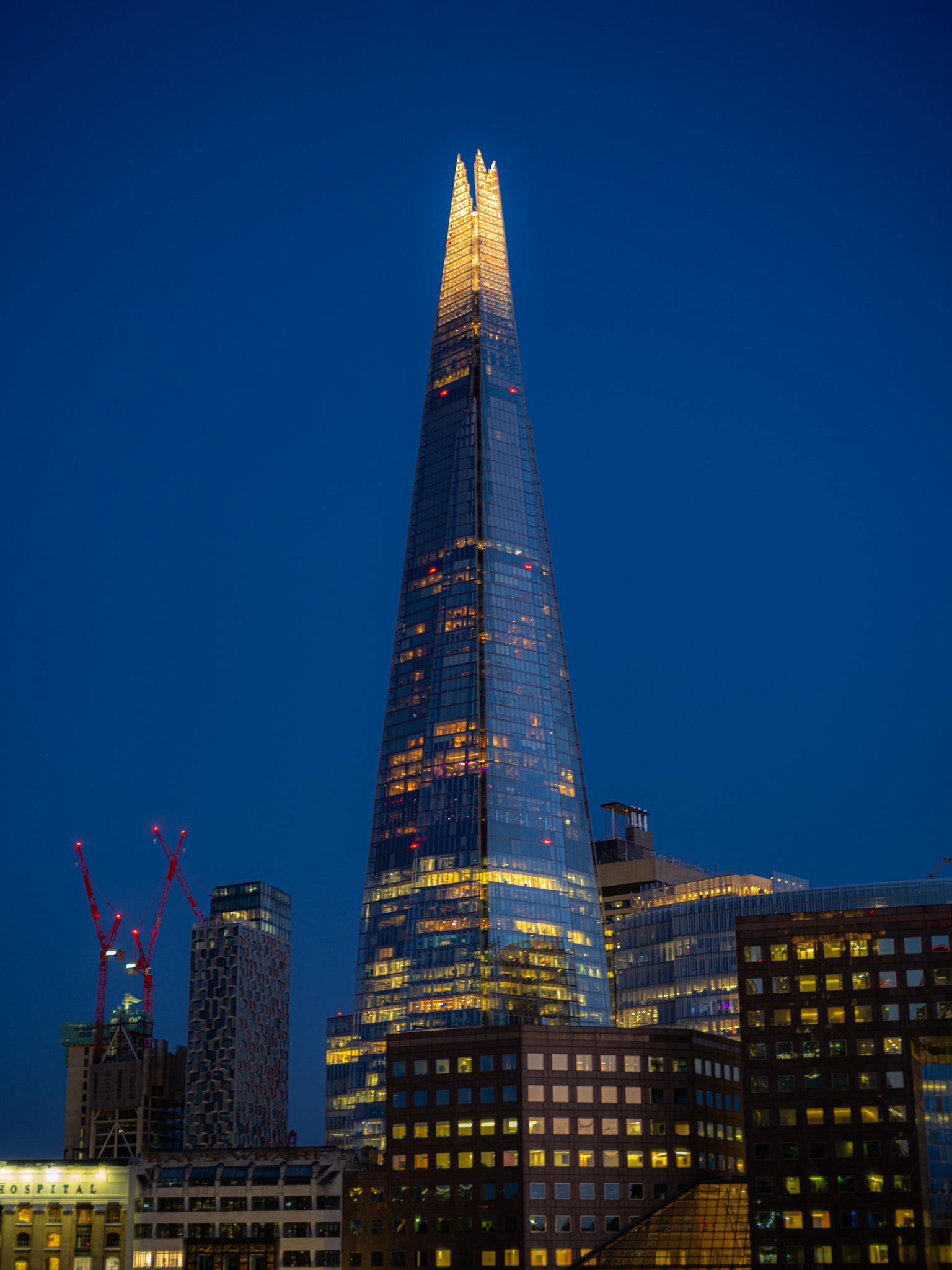 A vertical twilight photograph - by European elopement and wedding photographer Sturmsucht - of The Shard, London's tapering glass skyscraper, filling the centre of the frame against a clear, saturated dark-blue evening sky. The tower narrows to a shard-like point made of angled glass facets, and the last of the daylight or the building's lighting makes the upper section glow warm gold. Below that, scattered office lights — warm yellow and a few red aircraft warning lights — dot the mirrored facade, and the glass faintly reflects nearby buildings.

At the bottom of the frame sits a cluster of lower-rise buildings, their windows lit in warm yellow grids: a curved dark-brown office block in the centre foreground, a paler stone building on the left whose lit signage reads "…HOSPITAL" (the end of a word beginning off-frame), and more glass-fronted blocks on the right, one topped with rooftop plant or a small structure.

On the left, two red construction cranes stand silhouetted against the