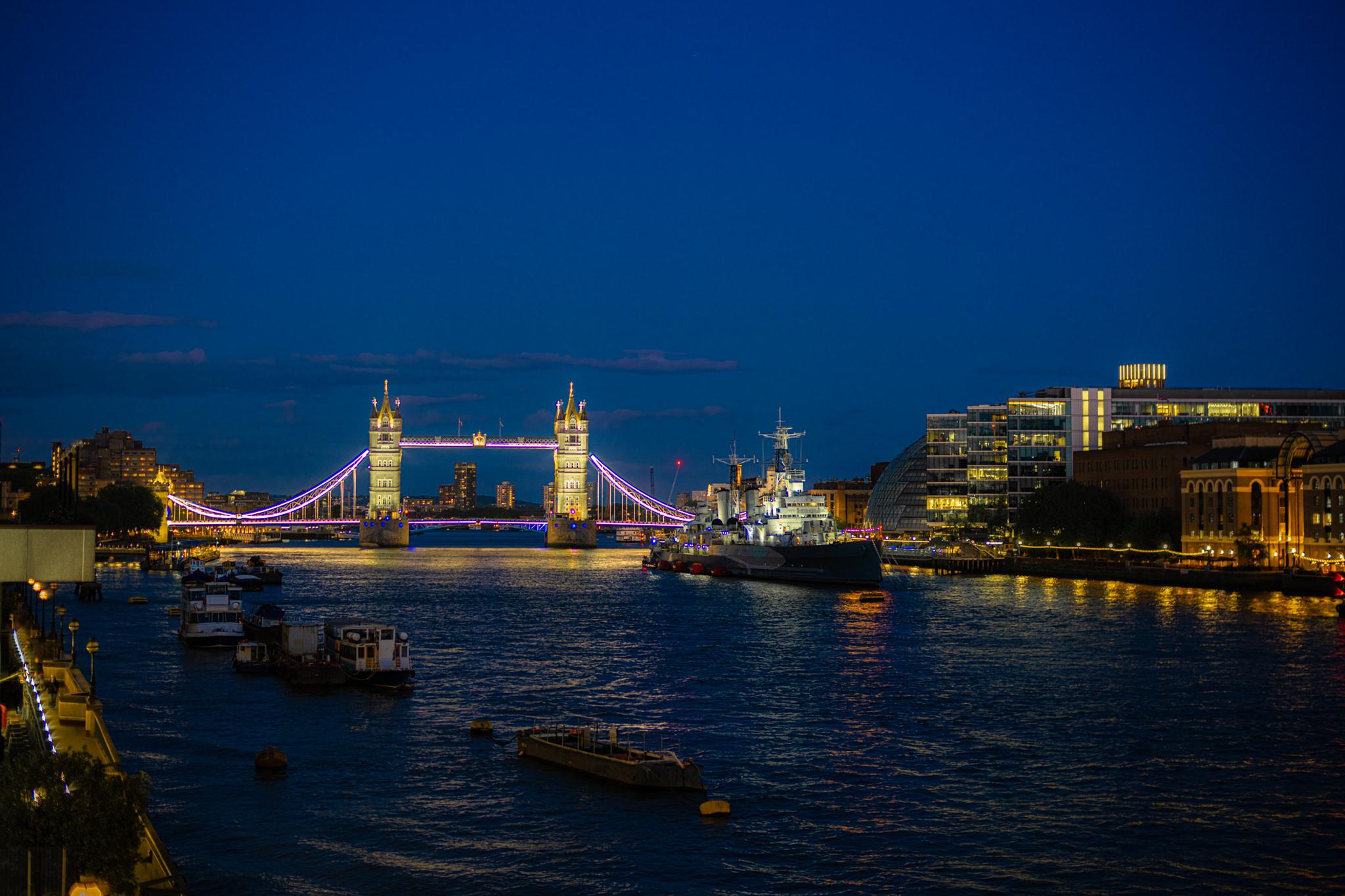 A wide twilight view of the River Thames in London under a deep blue evening sky with a few faint clouds low on the left horizon. The water fills the lower half of what is a landscape, 3:2 frame. A photograph by European elopement and wedding photographer Sturmsucht.

In the centre-left, Tower Bridge spans the river: two ornate stone towers joined by a high walkway, with the suspension chains sweeping down to either shore. The stonework is floodlit warm cream, while the cables and the lower deck carry bright violet-blue light. Warm reflections streak down the rippling water beneath it. Further bridges' lights and a lit skyline of blocks and towers recede behind the bridge on the left, where the moored HQS Wellington-style pier and riverside buildings sit.

To the right of the bridge, the grey Royal Navy cruiser HMS Belfast is moored mid-river, its masts, turrets and superstructure picked out in white light, with red buoys along the hull. Behind it, modern glass office blocks glow in gr