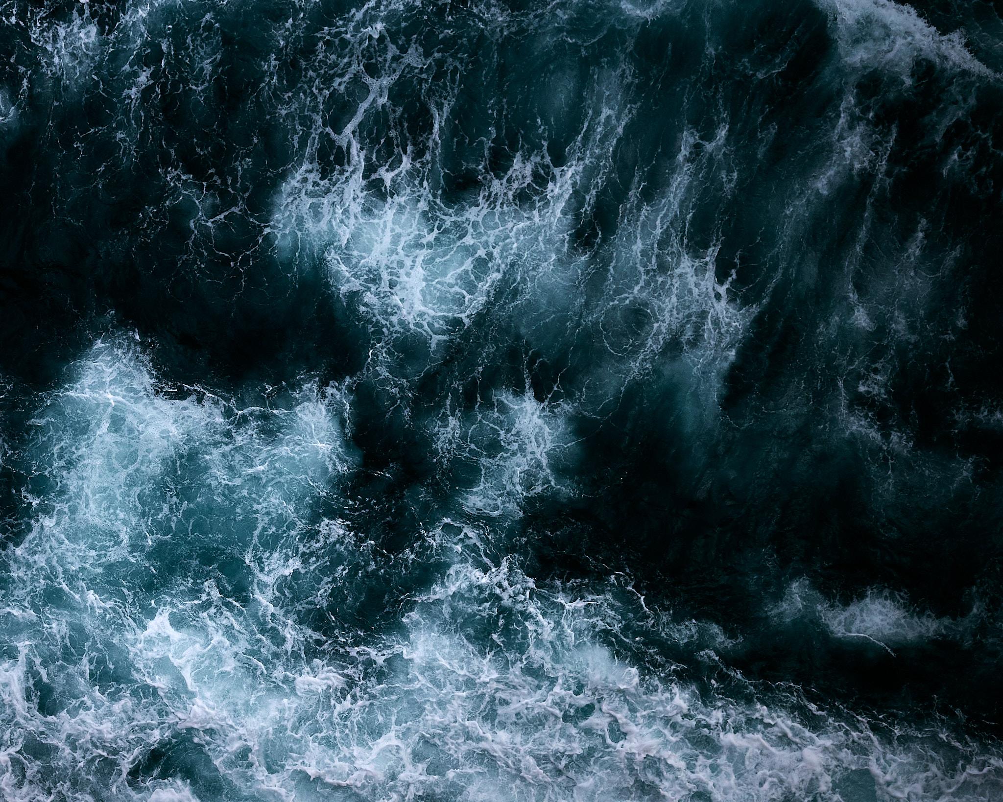 An aerial view of turbulent ocean water. The image captures dark blue and teal waves with white foam cresting at the surface. The composition emphasizes the movement and texture of the water, showcasing a dynamic interplay of light and shadow.
