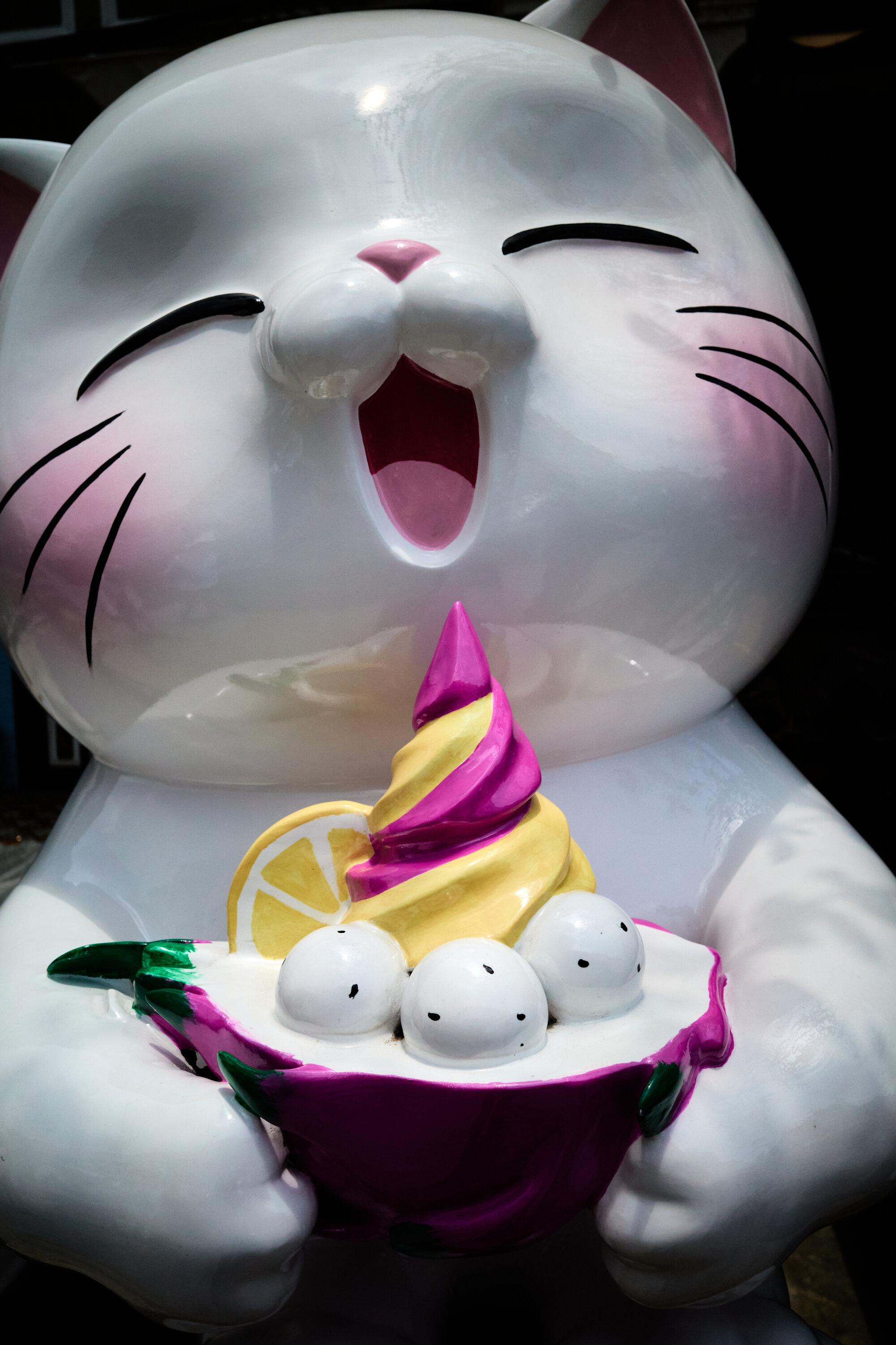 A large, whimsical statue of a cat with a joyful expression. The cat is holding a bowl of dessert featuring three white balls and a swirl of yellow and purple soft serve ice cream topped with a lemon slice. The statue is predominantly white with pink accents on the ears and nose.